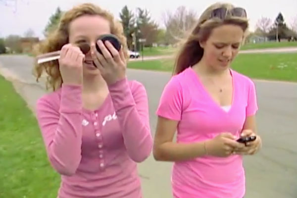 two girls walking. one putting on make-up, the other looking at her phone