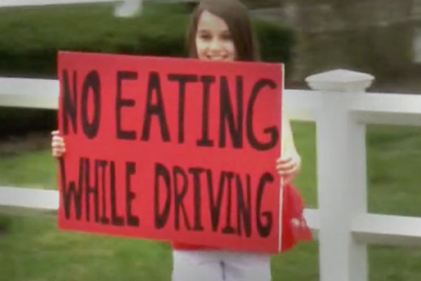 a young girl on the side of the road holding up a sign that says "no eating while driving"