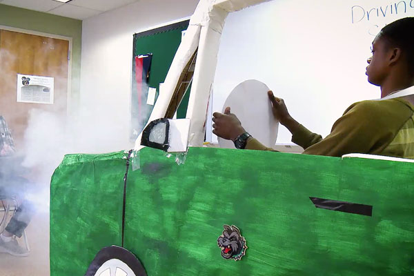 a boy driving in a green car made of cardboard with fog being blown at him